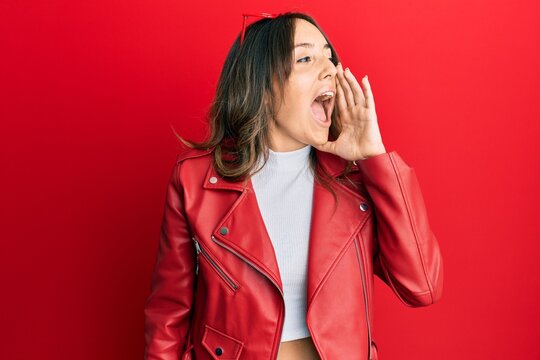 Young brunette woman wearing red leather jacket shouting and screaming loud to side with hand on mouth. communication concept.