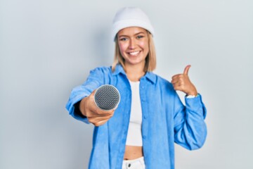 Beautiful blonde woman holding reporter microphone to the camera smiling happy and positive, thumb...