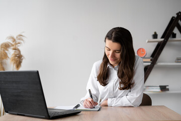 Young girl student studies at home online with a laptop while writing homework in a notebook while sitting at a desk. Remote work or education. lesson or conference via a laptop web.