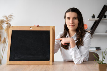 serious Caucasian teacher sitting in the classroom points his finger at the chalk board with space for text. Back to school concept. Millennial girl in the office holds a blackboard.