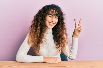 Young hispanic girl wearing casual clothes sitting on the table smiling with happy face winking at...