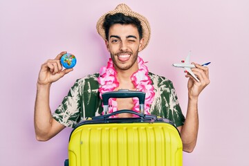 Young hispanic man wearing summer style and hawaiian lei holding world ball and plane toy sticking...