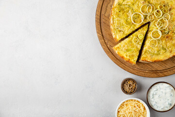 Quiche pie with leek, potatoes and cheese flat lay on gray concrete background with copy space and ingredients cheese, sour cream, onion rings on round wooden cutting board.