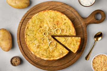 Quiche pie with leek, potatoes and cheese flat lay on gray concrete background and ingredients cheese, sour cream, onion rings on round wooden cutting board.