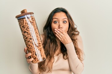 Young hispanic girl holding jar with macaroni pasta covering mouth with hand, shocked and afraid...