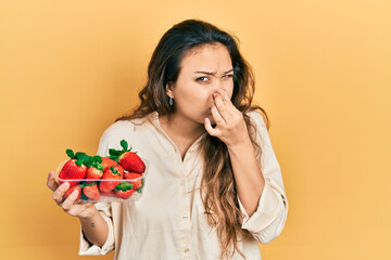 Young hispanic girl holding strawberries smelling something stinky and disgusting, intolerable...