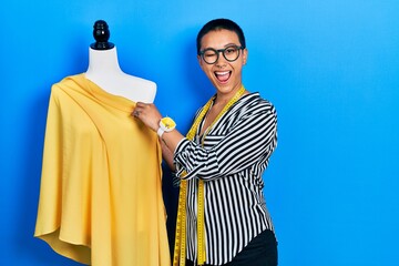 Beautiful hispanic woman with short hair standing by manikin sewing cloth winking looking at the...