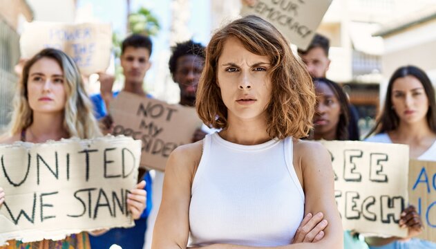 Young activist woman with arms crossed gesture standing with a group of protesters holding banner protesting at the city.