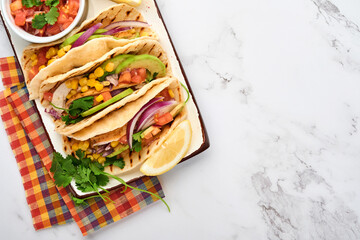 Mexican tacos with grilled chicken, avocado, corn kernels, tomato, onion, cilantro and salsa at white stone table. Traditional Mexican and Latin american street food. Top view.
