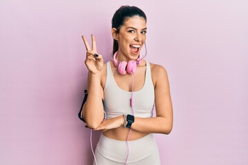Young hispanic woman wearing gym clothes and using headphones smiling with happy face winking at...
