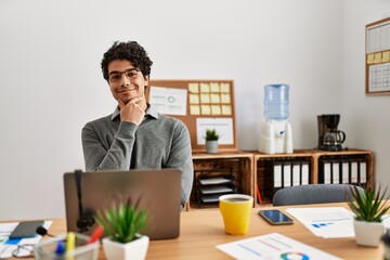 Young hispanic man wearing business style sitting on desk at office looking confident at the camera smiling with crossed arms and hand raised on chin. thinking positive.