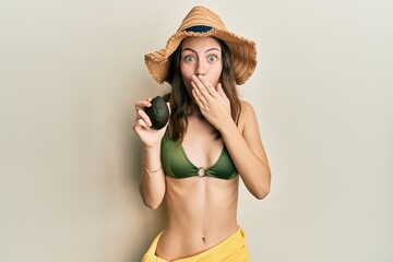 Young brunette woman holding avocado covering mouth with hand, shocked and afraid for mistake....