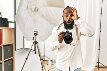 African american photographer man working at photography studio doing ok gesture shocked with surprised face, eye looking through fingers. unbelieving expression.