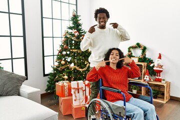 Young interracial couple with woman sitting on wheelchair by christmas tree smiling cheerful showing and pointing with fingers teeth and mouth. dental health concept.