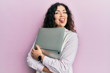 Young brunette woman with curly hair hugging laptop with love sticking tongue out happy with funny expression.