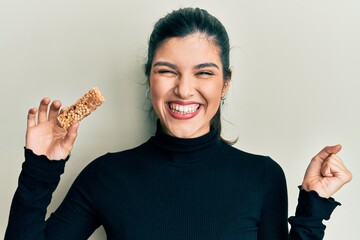 Young hispanic woman eating protein bar as healthy energy snack screaming proud, celebrating...
