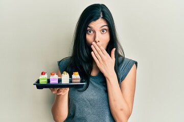 Young hispanic woman holding sweet pastries covering mouth with hand, shocked and afraid for...