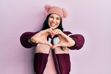 Young hispanic woman wearing wool winter sweater and cap smiling in love doing heart symbol shape with hands. romantic concept.