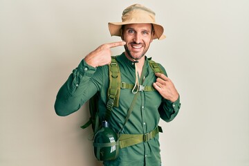 Handsome man with beard wearing explorer hat and backpack pointing with hand finger to face and nose, smiling cheerful. beauty concept