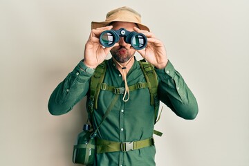 Handsome man with beard wearing explorer hat looking through binoculars looking at the camera blowing a kiss being lovely and sexy. love expression. - obrazy, fototapety, plakaty