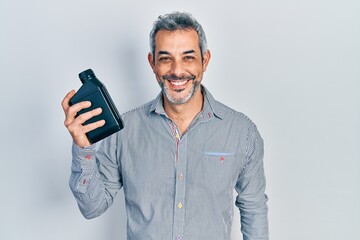 Handsome middle age man with grey hair holding motor oil bottle looking positive and happy standing and smiling with a confident smile showing teeth