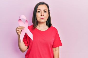 Middle age hispanic woman holding pink cancer ribbon thinking attitude and sober expression looking self confident