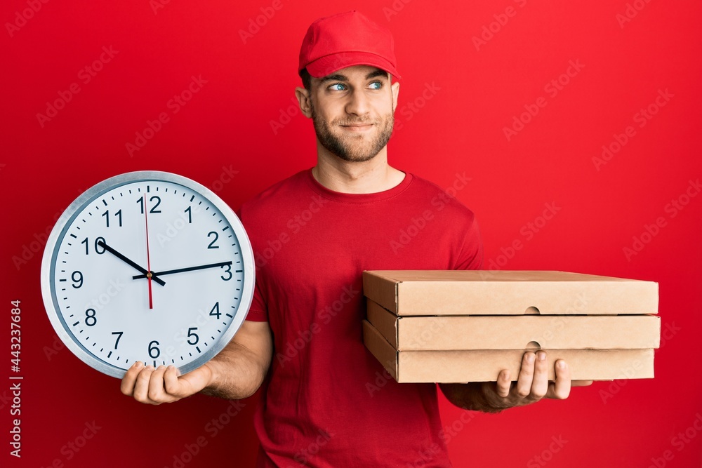 Sticker young caucasian man holding delivery package and clock smiling looking to the side and staring away 