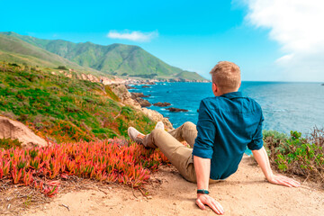 A young man is sitting on a high slope with a panoramic view of the picturesque ocean and mountains...