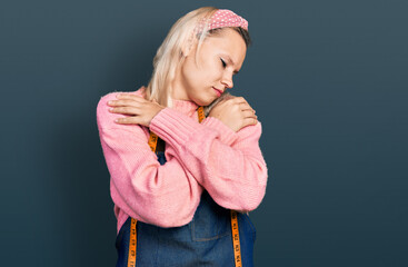 Young caucasian woman dressmaker designer wearing atelier apron hugging oneself happy and positive,...