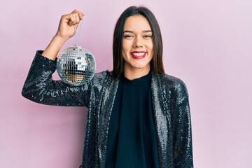 Young hispanic girl holding shiny disco ball looking positive and happy standing and smiling with a...