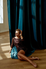 Woman in bronze dress posing on a wooden floor for fashion sitting in a dark room on a blue curtain background with shadow and rays of light on face - 443239021