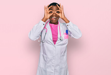 Young african american woman wearing doctor uniform and stethoscope doing ok gesture like binoculars sticking tongue out, eyes looking through fingers. crazy expression.