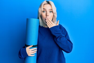 Young blonde girl holding yoga mat covering mouth with hand, shocked and afraid for mistake....