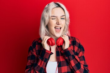 Young blonde girl listening to music using headphones winking looking at the camera with sexy...