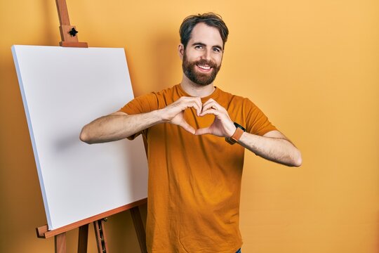 Caucasian man with beard standing by painter easel stand smiling in love doing heart symbol shape with hands. romantic concept.