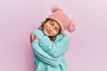 Little beautiful girl wearing wool sweater and cute winter hat hugging oneself happy and positive,...