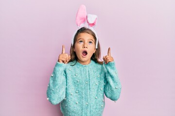Little beautiful girl wearing cute easter bunny ears amazed and surprised looking up and pointing...