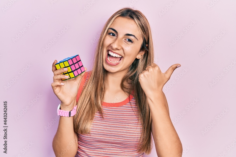 Sticker Beautiful hispanic woman holding colorful puzzle cube intelligence game pointing thumb up to the side smiling happy with open mouth