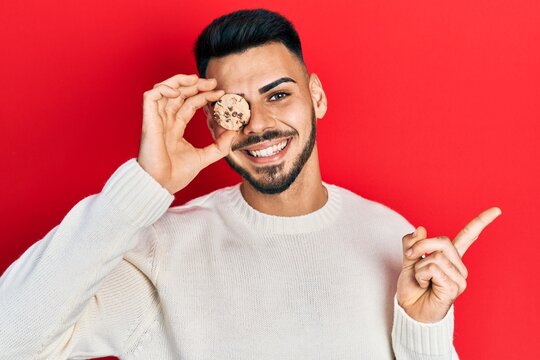 Young hispanic man with beard holding cookie over eye smiling happy pointing with hand and finger to the side