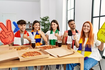 Group of young hispanic people eating pizza supporting soccer team at home smiling happy and positive, thumb up doing excellent and approval sign