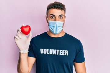 Hispanic young man wearing medical mask wearing volunteer t shirt holding heart afraid and shocked with surprise and amazed expression, fear and excited face.