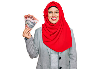 Beautiful hispanic woman wearing islamic hijab holding australian dollars banknotes looking positive and happy standing and smiling with a confident smile showing teeth