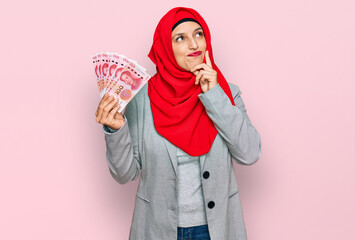 Beautiful hispanic woman wearing islamic hijab holding chinese yuan banknotes serious face thinking about question with hand on chin, thoughtful about confusing idea