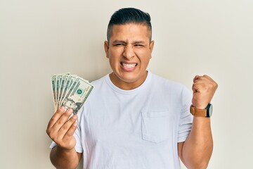 Young latin man holding 20 dollars banknotes screaming proud, celebrating victory and success very excited with raised arm