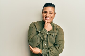 Fototapeta premium Young latin man wearing casual clothes looking confident at the camera smiling with crossed arms and hand raised on chin. thinking positive.