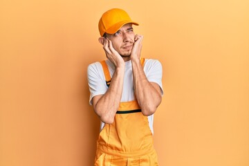 Hispanic young man wearing handyman uniform covering ears with fingers with annoyed expression for the noise of loud music. deaf concept.