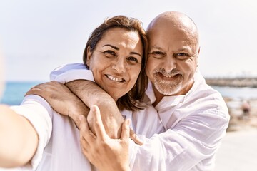 Middle age hispanic couple of husband and wife together walking by the beach on a sunny day. Hugging in love on vacation to the seaside taking a selfie picture