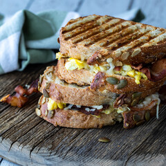 Two grilled breakfast paninis with egg and bacon on a rustic wooden board.