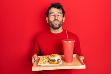 Handsome man with beard eating a tasty classic burger with fries and soda looking at the camera blowing a kiss being lovely and sexy. love expression.