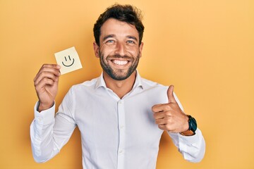 Handsome man with beard holding smile emoji reminder smiling happy and positive, thumb up doing...